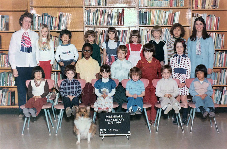 Color photograph of Mrs. Hudgins’ class during the 1975 to 1976 school year. The children appear to be in first or second grade. 17 children are pictured, seven of whom are girls. Principal Calvert is standing on the left and Mrs. Hudgins is standing on the right of the children. The children are arranged in three rows with one row seated and two rows standing. In front of the seated children is a sign that reads: Forestdale Elementary, 1975 – 1976, Miss Calvert, principal. A Sheltie breed dog is sitting on the floor next to this sign. 