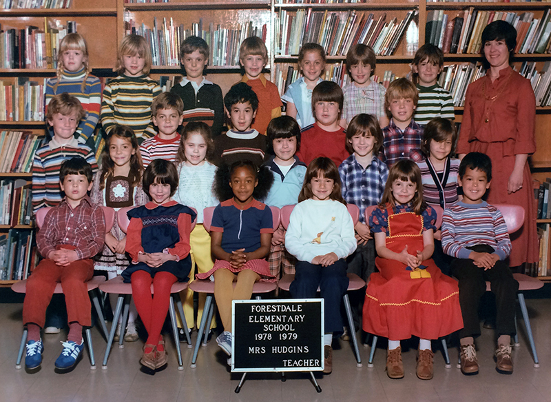Color photograph showing Mrs. Hudgins’ class at Forestdale. 23 children are pictured, arranged in four rows. Hudgins can be seen on the far right behind her students. She is wearing a red blouse and skirt. 