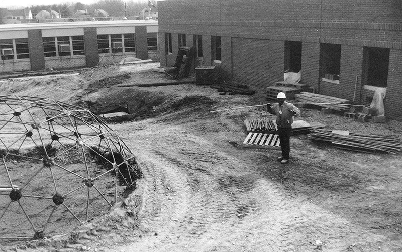 Black and white photograph of construction progress on the music room and art classroom addition to Forestdale. The picture is from our 1992 to 1993 yearbook. Window-mounted air conditioning units are visible in the classroom windows. A construction worker, wearing a hard hat, is carrying supplies on his shoulder. The new classroom wing is visible behind him. It is two-stories tall and has a brick veneer. The windows are not yet in place, and construction materials are placed around the structure.