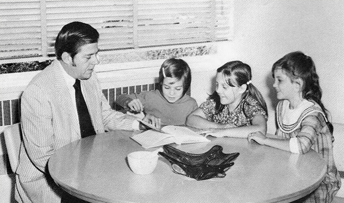 Black and white yearbook portrait of Principal Ronald West. This picture comes from a 1971 to 1972 yearbook at North Springfield Elementary School where West became principal after leaving Forestdale. He is seated at a small round table reading a book to three students. 