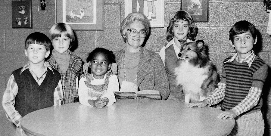 Black and white portrait of Principal Nancy Calvert from Forestdale’s 1977 to 1978 yearbook. She is seated at a table surrounded by five students. Dutchess is seated on a stool between two students to the right of Calvert.  