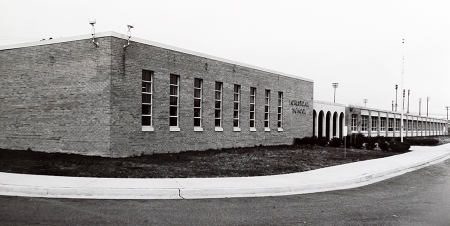 Black and white photograph of the front of Forestdale Elementary School taken in the late 1960s. The photograph was taken from the south end of the bus loop and shows the cafeteria in the foreground, main entrance in the mid-ground, and the front classroom wing. The lights of the Lee High School stadium are visible in the far distance. There are shrubs planted in front of the school.